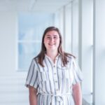 Een glimlachende vrouw in een lichtgekleurde, gestreepte jurk poseert in een moderne kantooromgeving met grote ramen en natuurlijk licht.