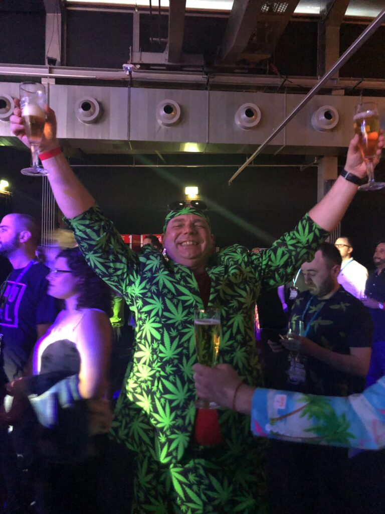Verjaardagsviering met feestende man in kleding met marijuana blad print, proostend met een glas bier op een druk evenement met verlichte sfeer.