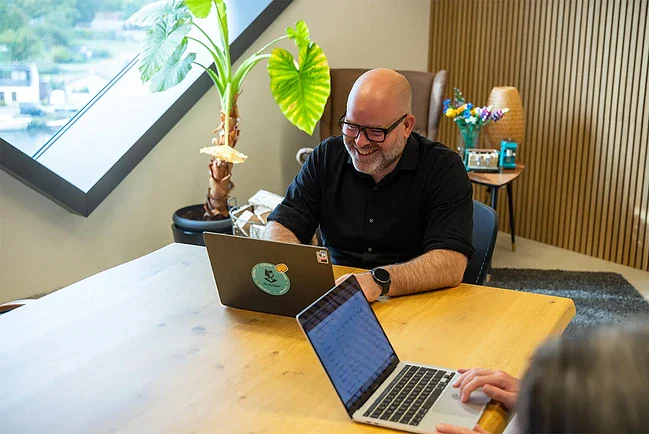 Een collega zit aan tafel in de vergaderruimte en lacht achter zijn laptop.