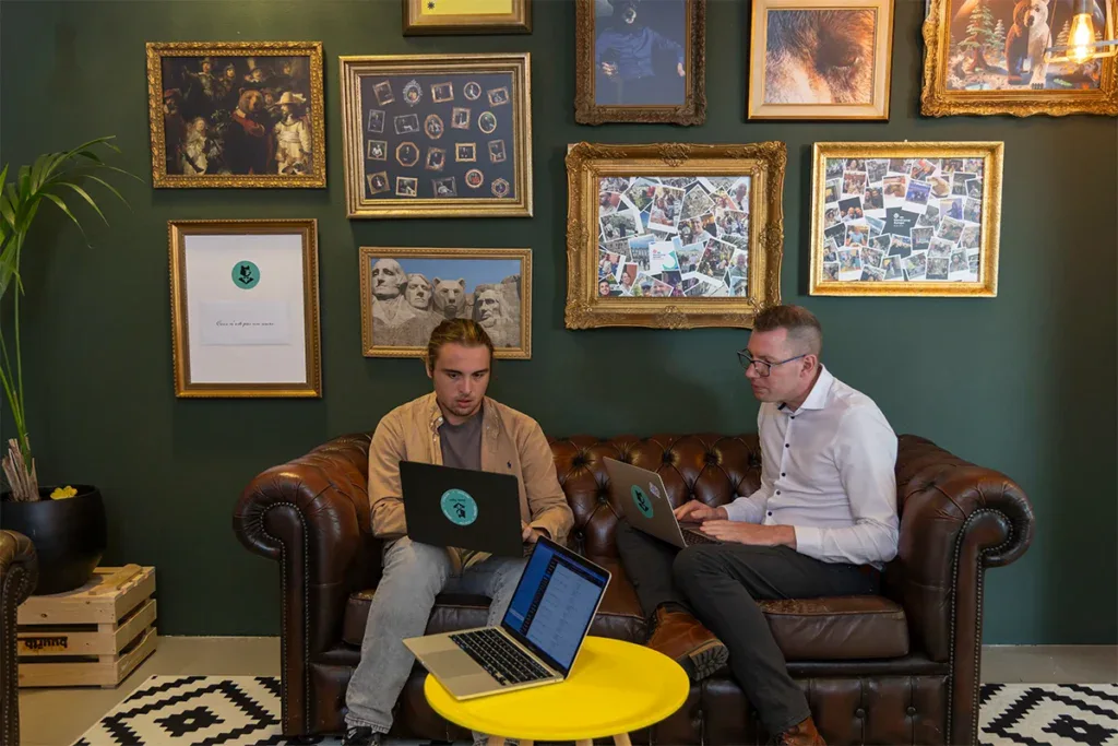 Twee developers zitten op een bank met een laptop op schoot en zijn aan het overleggen.