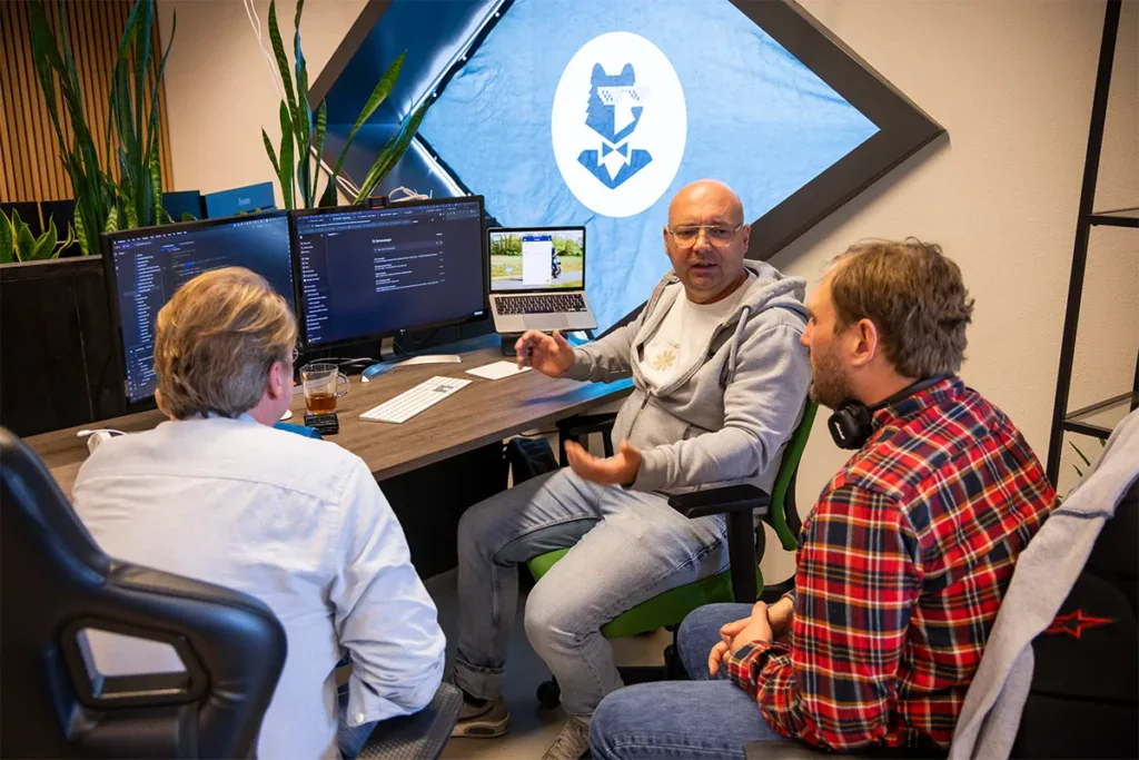 Drie programmeurs zijn aan het overleggen en zitten bij een bureau met beeldschermen.