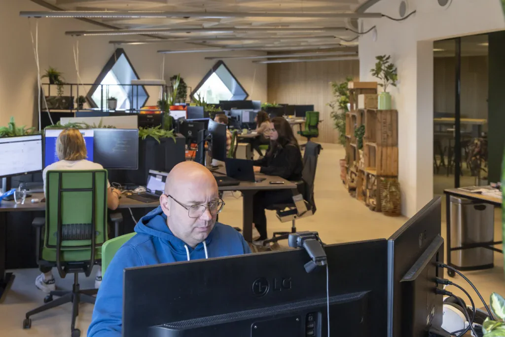 Back end developer aan het werk achter zijn bureau op kantoor met in de achtergrond collega's die ook aan het werk zijn.