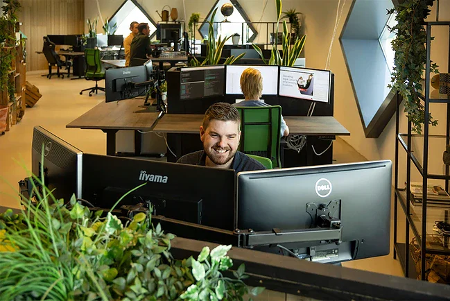 Front end developer zit lachend achter zijn bureau te werken. In de achtergrond zie je collega's aan het werk.
