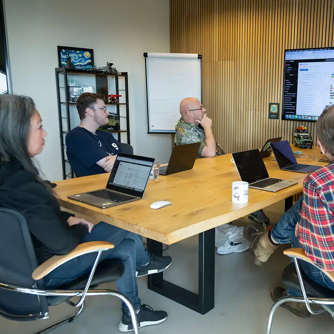 Het team zit aan tafel en kijkt naar een scherm tijdens de teammeeting in de boardroom.