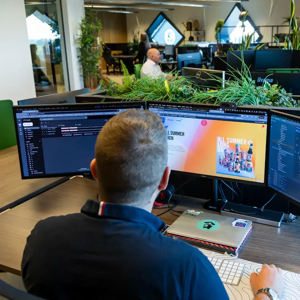 Front end developer werkt achter zijn bureau aan en website op kantoor, in de achtergrond zie je collega die ook aan het werk zijn.