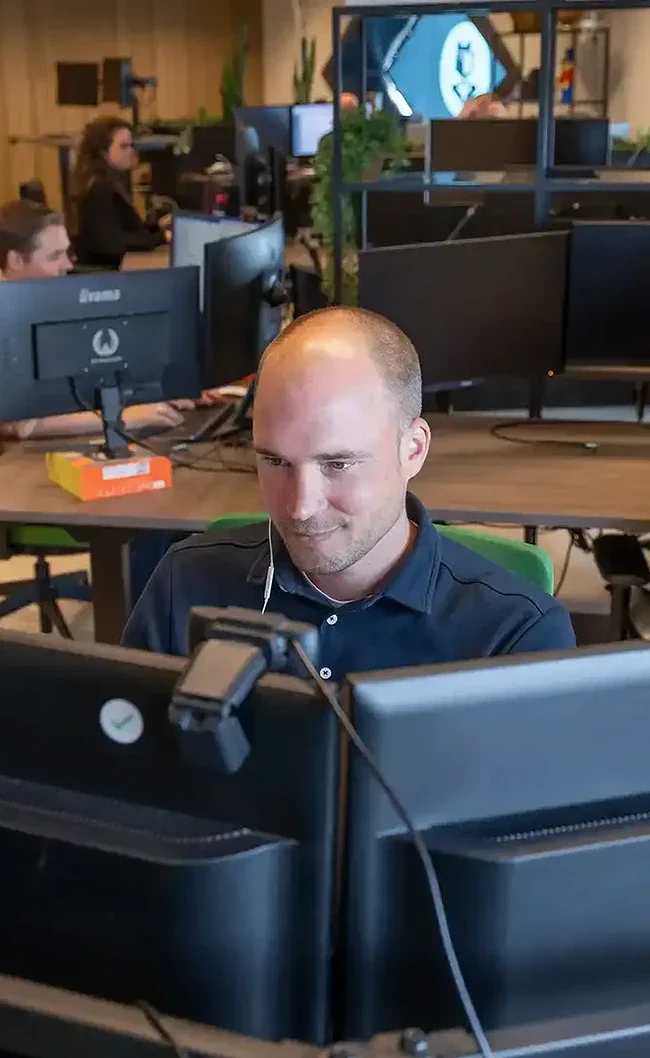 Front end developer zit achter zijn bureau te programmeren, op de achtergrond zie je collega's aan het werk.